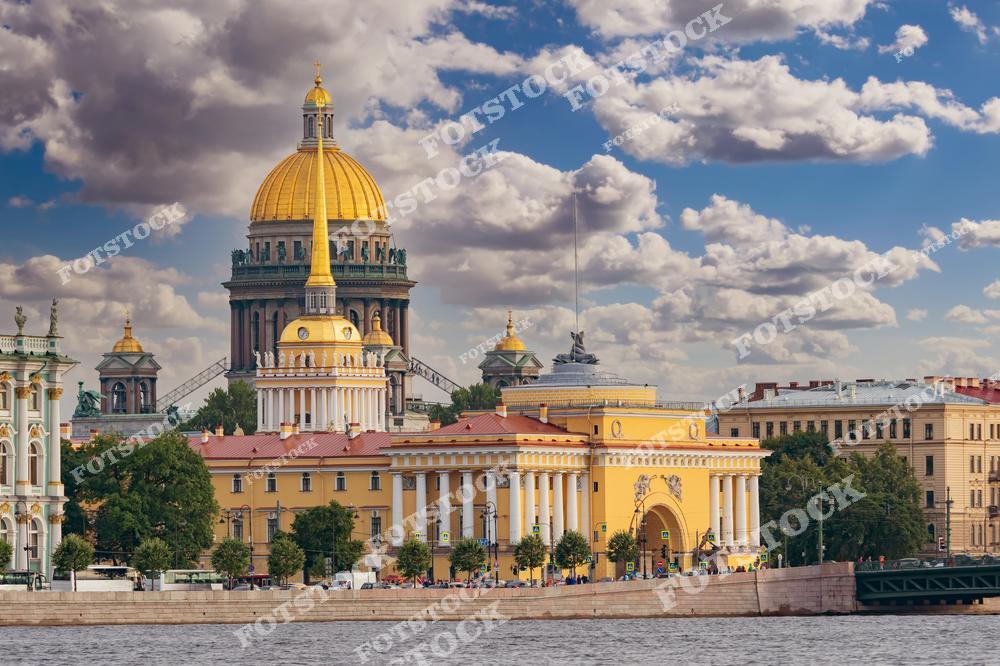 На снимке изображены знаменитые достопримечательности Санкт-Петербурга — здание Адмиралтейства и Исаакиевский собор. Золотой шпиль Адмиралтейства и купол собора выделяются на фоне голубого неба с облаками. Вид с Невы подчеркивает величие архитектурного ансамбля, сочетающего элементы классического стиля и барокко. Это место является знаковым для города и популярно среди туристов.